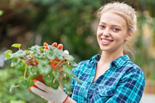 A young woman with blonde hair pulled back, dressed in a blue and black plaid shirt, is smiling and holding a small potted plant with green, rounded leaves and tiny white flowers. She is standing outdoors in a garden setting with blurred greenery in the background, suggesting a lush, well-maintained outdoor space typical of a domestic garden within Uxbridge. The scene indicates activities related to gardening or plant care, aligning with professional gardening services offered by Landscapers Uxbridge. The natural outdoor environment features a mix of foliage with soft natural light, highlighting the vibrant green tones of the plants and the friendly, attentive attitude of the gardener, exemplifying quality lawn and garden care in the local area.