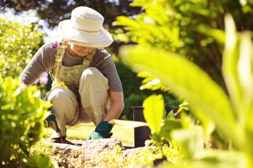 A gardener wearing a wide-brimmed white hat, beige overalls, and green gardening gloves is kneeling on a well-maintained lawn in a garden, tending to a flower bed bordered by soil and decorative paving stones. The garden features lush green bushes, shrubs, and mature trees with dense foliage, creating a vibrant outdoor environment. Bright sunlight illuminates the scene, casting natural light across the grass and plants, indicating a clear, warm day. In the background, there are additional garden elements such as potted plants and natural wooden fencing, suggesting a landscaped, residential garden space typical of Uxbridge. This scene reflects professional gardening and landscaping activities that a company like Landscapers Uxbridge might undertake in local gardens, focusing on maintenance, planting, and outdoor care services.