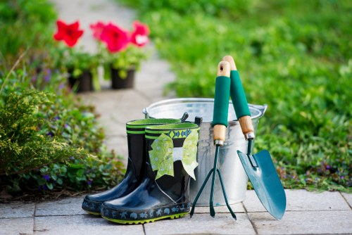 The image shows a garden scene featuring a pair of black rubber gardening boots placed on a paved pathway, with a pair of green-handled garden tools—possibly a trowel and a hand fork—resting inside a white plastic bucket nearby. In the background, there are vibrant pink and red flowering plants, with some potted flowers visible at the edge of a well-maintained grassy lawn area. The lush green foliage and the neatly bordered flower bed suggest a well-tended outdoor space, typical of a residential garden in Uxbridge, with natural daylight illuminating the scene. This setup reflects a typical gardening or landscaping environment, suitable for outdoor maintenance or planting activities, and demonstrates natural textures such as soil, paving, metal tools, and plant foliage, consistent with professional gardening services provided by Landscapers Uxbridge.