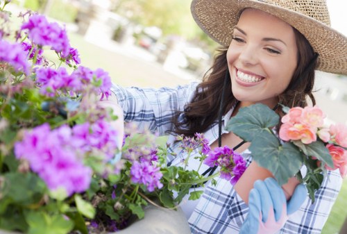 A woman with long brown hair, wearing a straw hat, a light blue and white checkered shirt, and blue gardening gloves, is tending to a variety of flowering plants in a garden. She is smiling while carefully handling vibrant purple and pink blossoms, which are surrounded by lush green foliage. The garden features a mix of flower beds with blooming plants, neatly maintained borders, and a lawn area in the background. The scene is outdoors on a sunny day, with natural light illuminating the colourful flowers and greenery. This landscaping and gardening activity reflects typical garden maintenance practices, possibly carried out by specialists such as Landscapers Uxbridge, to enhance outdoor spaces in the Uxbridge area, near postcodes UB8 or UB9, supporting garden renovation and outdoor beautification services.