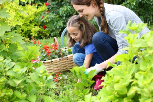 A woman and two young girls are working together in a lush, well-maintained garden in Uxbridge, surrounded by vibrant green foliage and colorful flowering plants. The women and children are crouching down near a low hedge of leafy bushes, with the woman guiding the children as they place small red flowers or seedlings into the soil. Behind them, a curved flower bed filled with soil and blossoming plants borders a neatly mown lawn that extends towards a backdrop of mature trees and shrubs, providing privacy and shade. The garden features a combination of different textures, including grassy areas, smooth paving stones, and wooden decking visible in the background, creating an inviting outdoor space suitable for gardening activities and relaxation. The weather appears bright and clear, with natural sunlight illuminating the scene, emphasizing the vibrant greens and natural tones of the garden's plants and surfaces. The image reflects a typical landscaped outdoor area in Uxbridge, representative of local gardening services offered by landscapers such as Landscapers Uxbridge, who specialise in garden makeovers and outdoor maintenance tasks to enhance residential gardens in the area.