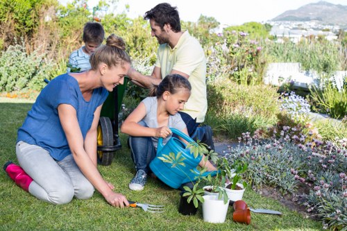 A family working together in a well-maintained residential garden in Uxbridge, with lush green grass, flower beds, and a variety of seasonal plants and shrubs. The foreground features a woman kneeling on the lawn, planting or tending to a small shrub, while a young girl with a blue backpack assisting nearby, surrounded by gardening tools and plant pots. In the background, a man and another child are sitting on the lawn, engaging in outdoor activities amidst an environment characterized by natural blooming flowers, decorative foliage, and a tidy lawn area. The garden is set under bright, clear weather, highlighting the natural tones of the green grass, colorful blossoms, and aged wooden borders that define different planting zones, representing a typical outdoor space that benefits from professional gardening services such as lawn care, planting, and landscaping in the Uxbridge area managed by Landscapers Uxbridge.