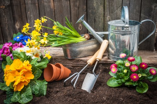 A close-up view of a well-maintained garden corner featuring freshly turned dark soil with various gardening tools and colourful potted plants. The soil area is bordered by a weathered wooden fence in the background, providing a natural enclosure typical of a Uxbridge backyard. On the soil, there is a metal watering can with a long spout, a pair of small hand tools—a trowel and a hand fork with light wooden handles—and an empty terracotta pot. To the left, vibrant primroses with bright yellow, pink, and purple flowers sit among lush green leaves, while a plant with pink and white daisy-like flowers is positioned on the right side. Additionally, a small gardener's rake rests on the soil, ready for recent planting or soil aeration. The overall scene reflects ongoing gardening activity, with fresh flowers and gardening implements indicating maintenance work or planting efforts in a typical residential outdoor space, with natural daylight highlighting the vivid colours and textures appropriate for garden and landscaping services in Uxbridge.