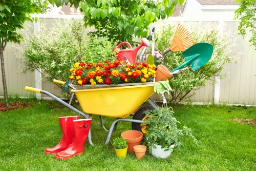 A colourful garden scene in a residential outdoor space featuring a yellow wheelbarrow filled with vibrant red and yellow marigold flowers, surrounded by gardening tools and accessories including a red watering can, a pair of red rubber boots, and various small flower pots placed on a well-maintained green lawn. In the background, there are lush bushes, small trees, and a white fence, with some shade provided by overhanging tree branches. The overall environment appears bright and sunny, suggesting clear weather, and the setting reflects a well-kept garden prepared for planting and outdoor maintenance, typical of Uxbridge gardens managed by landscapers or gardening service providers like Landscapers Uxbridge.