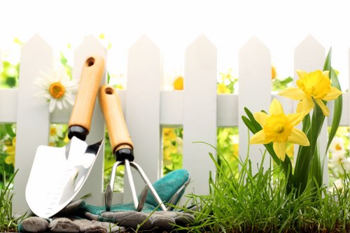 In a well-maintained garden, a white wooden picket fence runs across the background, creating a defined boundary for a landscaped outdoor space. In the foreground, there is a patch of lush, green grass with a dense, healthy appearance. To the right, a cluster of vibrant yellow daffodils with green foliage adds bright natural colour to the scene. The garden contains a mixture of soil and planted flower borders, and there are gardening tools, including a small trowel and a hand rake, leaning against the fence, indicating recent or ongoing gardening work. The scene is well-lit, suggesting a bright, sunny day, which highlights the vivid colours and textures of the grass, flowers, and tools. The overall environment appears orderly and cared for, matching the services offered by companies like Landscapers Uxbridge that specialise in outdoor gardening and landscaping maintenance in the local area, such as UB8 postcode or nearby locations in Uxbridge. This image demonstrates a typical small garden space suitable for lawn care, planting, and general outdoor enhancement, reflecting attention to detail in landscape design and maintenance.
