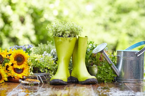 The image displays a gardening scene set outdoors in a garden area with a natural, lush green background. In the foreground, there is a wooden surface, likely a garden table or workbench, showing visible wood grain. Positioned centrally are a pair of bright green rubber gardening boots filled with small white flowering plants, with the boots standing upright and slightly apart. To the left of the boots, there is a bouquet of sunflowers and other mixed flowers with yellow, purple, and white blooms. To the right of the boots, there is a metallic watering can and a small gardening trowel with a wooden handle, both placed on the wooden surface. The scene suggests a typical outdoor gardening environment, highlighting tools and plants commonly used in garden maintenance and landscaping work, and it subtly supports local gardening and landscaping services in Uxbridge. The lighting indicates daytime with natural sunlight illuminating the scene, emphasizing the freshness and vibrancy of the plants and tools.