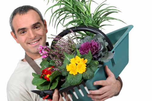 The image depicts a man with short dark hair and a friendly expression, holding a plastic garden watering can filled with a variety of vibrant flowering plants, including yellow, purple, and red blossoms, along with lush green foliage. The background suggests an outdoor garden environment with local Uxbridge scenery, featuring a neatly maintained lawn or grassy area, bordered by shrubbery or hedges. In the foreground, a section of the garden with fresh soil and possible paving or decking elements can be observed, indicating an outdoor space prepared for gardening or landscaping activities. The overall scene conveys a sense of nurturing and outdoor gardening care, which aligns with services offered by landscapers in Uxbridge, such as lawn maintenance, planting, and garden enhancements. The weather appears to be bright and clear, emphasizing natural sunlight that highlights the vivid colours and healthy appearance of the plants, suitable for professional landscaping and gardening work by Landscapers Uxbridge.