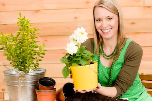A woman with shoulder-length blonde hair smiling while sitting outdoors in a garden setting, holding a yellow flower pot containing a green leafy plant with white flowers. In front of her on a wooden surface, there is a small green shrub in a metallic pot, a black plant pot, and an empty reddish-brown pot. The background features a light wooden fence, and the overall scene is well-lit with natural daylight, suggesting a bright, pleasant day. The garden area appears well-maintained, with a variety of potted plants and gardening containers, indicative of outdoor gardening activities typical in residential outdoor spaces in Uxbridge or nearby areas. The scene emphasizes gardening care and outdoor plant cultivation, aligning with professional garden and lawn services offered by Landscapers Uxbridge.