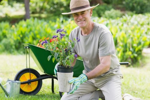 A middle-aged man wearing a straw hat, light grey t-shirt, and gardening gloves is kneeling on a well-maintained lawn in a garden, smiling while holding a pot filled with purple and red flowering plants. Behind him, there is a green wheelbarrow with a yellow wheel, partially filled with gardening tools and additional plants, set on a lush, green lawn. The garden features dense hedges, shrubs, and a mixture of small flowering plants, with a background of tall trees and shadowed areas under a bright, clear sky, indicating a sunny day. The overall scene depicts a neatly landscaped outdoor space suitable for gardening and outdoor maintenance, reflecting professional gardening services in Uxbridge.