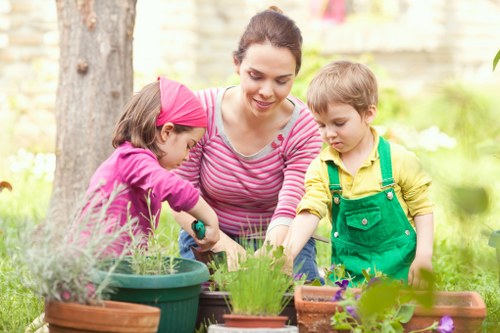 A woman and two children are engaged in gardening activities in a well-maintained backyard garden, with a mature tree trunk visible on the left and a background of blurred green foliage. The woman, wearing a pink and white striped top, is smiling as she assists the children with planting or handling small plants. The girl, dressed in a pink headband and matching pink long-sleeved top, is focused on a plant in a large green pot, while the boy, in a yellow shirt with green overalls, is also participating in the gardening activity. The garden features a variety of small potted plants, including flowering and foliage species, placed on a surface that may be a garden table or ground, with some in rectangular containers and others in round pots. The scene is set during daylight hours with natural sunlight illuminating the outdoor space, emphasizing the lush greenery, vibrant plant life, and the natural textures of soil, pots, and garden surfaces, illustrating a peaceful and productive gardening environment typical of Uxbridge's outdoor spaces.